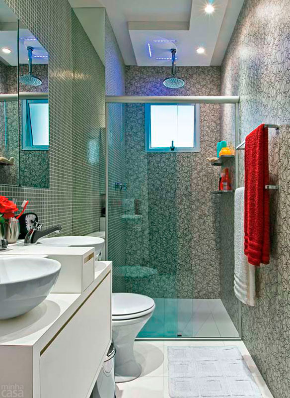 Um laminado decorativo com estampa abstrata reveste a parede do boxe deste banheiro projetado por Ricardo Umada. Na outra parede, ele optou por pastilhas de vidro em tom neutro. As fitas de led ao redor da ducha mudam de cor e permitem diferentes ambienta&ccedil;&otilde;es. Os acess&oacute;rios de tom prateado harmonizam com o cinza predominante.