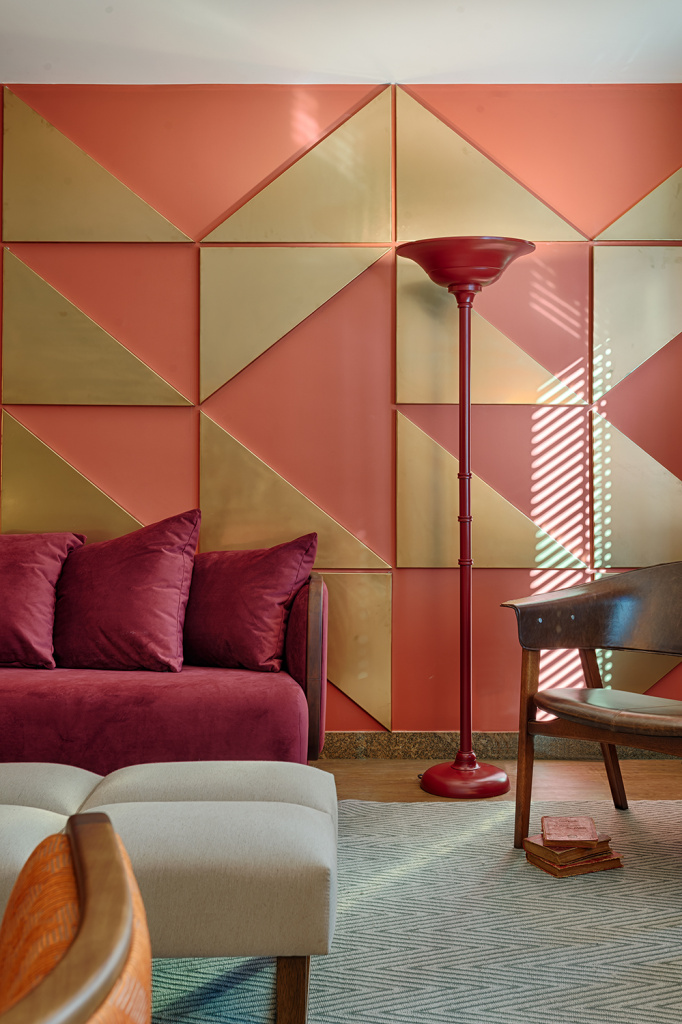 Esta sala, criada pela Melina Mundim, trabalha principalmente com tons terrosos e avermelhados, com exce&ccedil;&atilde;o das cadeiras ao fundo, dos bancos, do tapete e dos pain&eacute;is triangulares. A decora&ccedil;&atilde;o tamb&eacute;m possui uma influ&ecirc;ncia retr&ocirc;, como pode ser observado na lumin&aacute;ria de ch&atilde;o vermelha, nos formatos arredondados do sof&aacute; e das poltronas e na estampa geom&eacute;trica criada pelos pain&eacute;is dourados na parede.