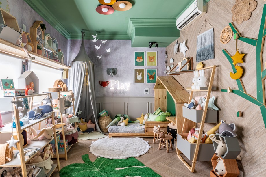 Espaço Conceito - Paula Carvalho. Pensado como um quarto para encantar crianças de todas as idades, este espaço funciona como uma loja e reúne produtos de seis marcas de decoração. Uma boiserie forra parte das paredes, o mesmo ocorre com um painel adesivo de estampa botânica. Dispostos por todo o ambiente, cestos e almofadas apresentam texturas aconchegantes e divertidas. Destaque para a cama casinha.