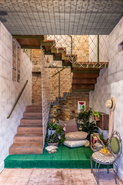 Hall de entrada e Bilheteria - Isabela Scaff e Maria Gabriela Azevedo, e paisagismo de Clariça Lima. Com 15 m² e pé-direito duplo, o hall é aberto para os dois andares da casa. O rústico ganha espaço na ambientação, como nas cordas que entrelaçam o guarda-corpo original em ferro. No vão, o mobiliário convive com plantas e flores, além do tapete que imita grama.