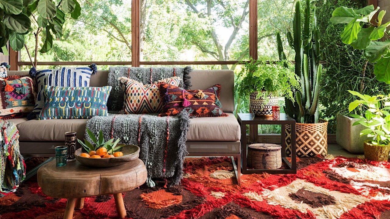 Decoraçnao estilo boho com tapete vermelho estamapado, sofá cinza com almofadas estampadas coloridas e muitas plantas