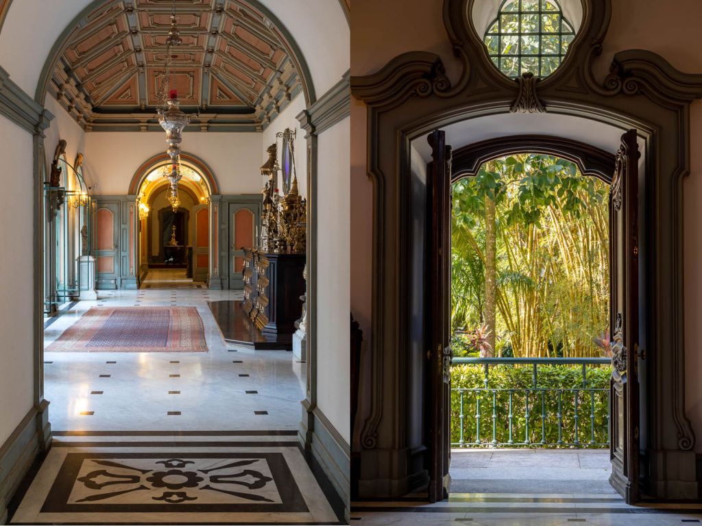 À esquerda, corredor com piso com mosaicos, teto adornado e móveis em madeira com ornamentos. À direita, porta em madeira com ornamentos que se abre para um jardim verde