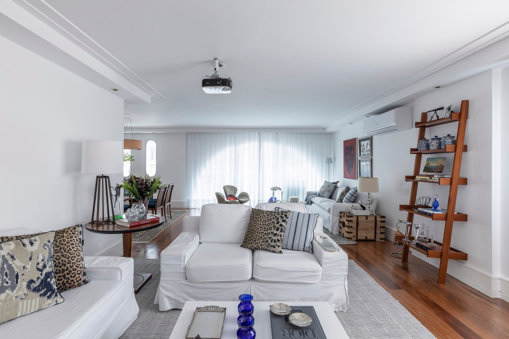 Sala de estar com sofás e paredes brancas, com forro em gesso nas laterais por conta da iluminação. No chão de madeira, uma tapete cinza decora o espaço, assim como almofadas estampadas. Um mesa de centro, de lateral e uma estante completam o ambiente