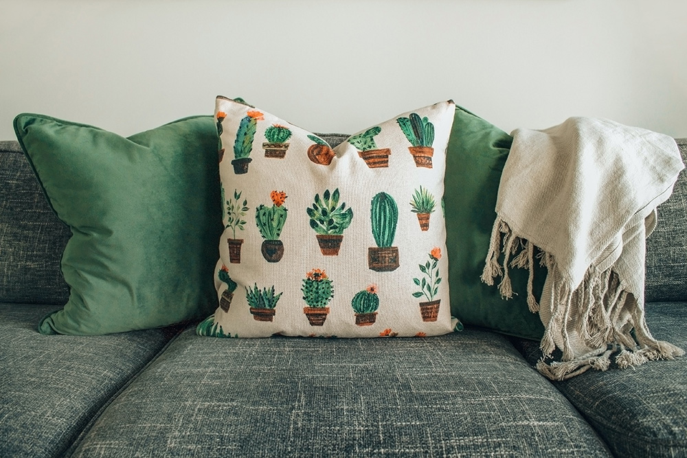 Sofá verde com três travesseiros: dois verdes lisos e um branco com estampa de cactus.