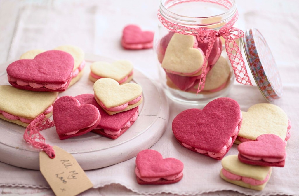 Biscoitos em formato de coração amarelos e rosas. Pote de vidro com fita rosa e mais biscoitos dentro ao fundo