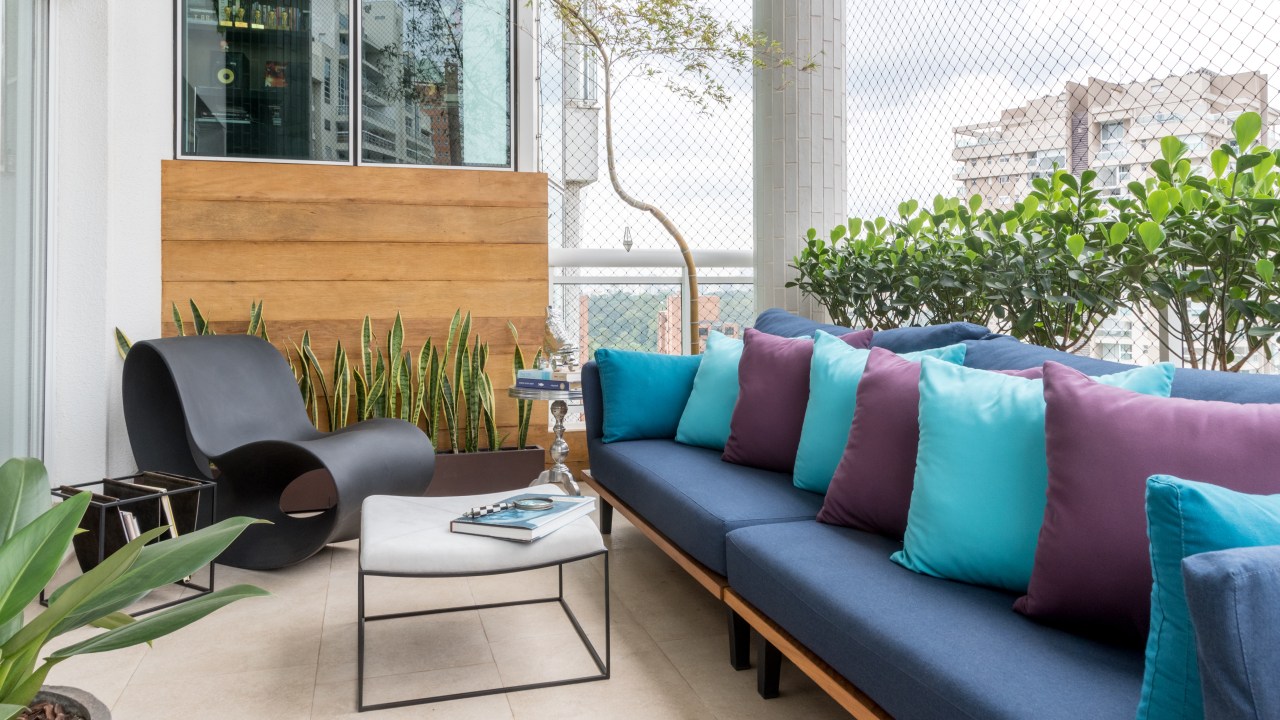 Varanda aberta com porcelanato, mesa de centro branca, com sofá azul com almofadas em azul claro e roxo. Ao lado, com plantas ao fundo, uma cadeira de design completa o ambiente