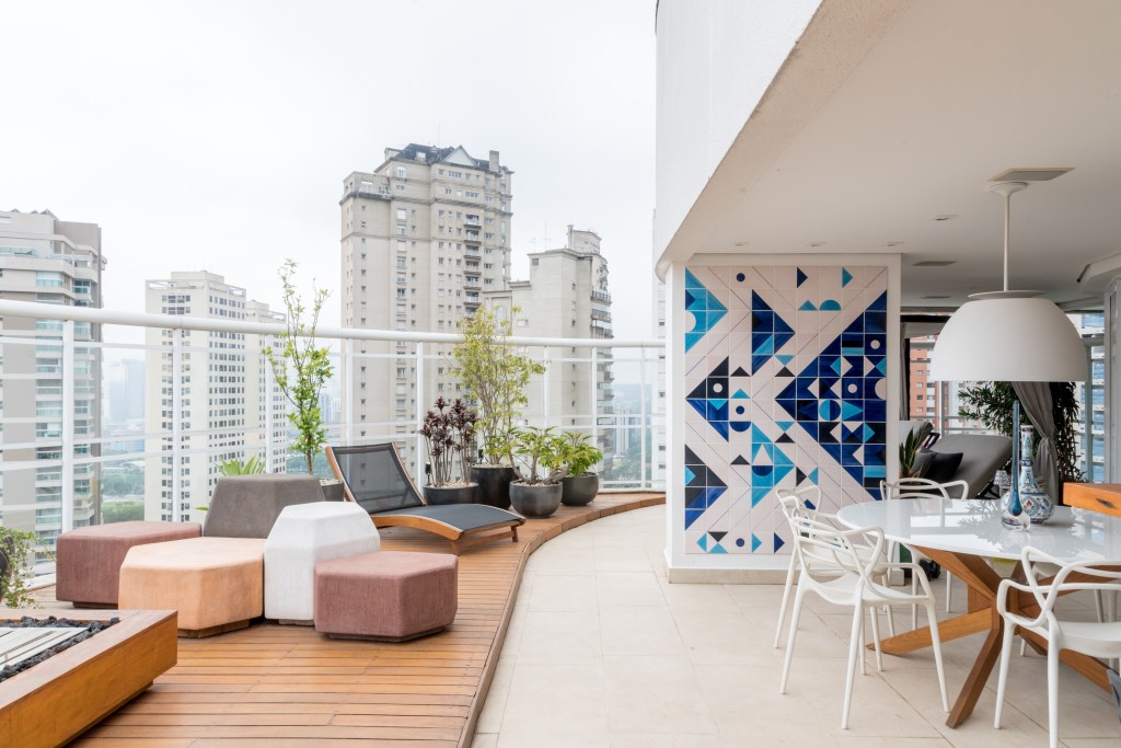 Varanda da cobertura com madeira com proteção para o deck inserido na área externa, combinada com o porcelanato. O mobiliário é composto por puffs em tons de terracota e salmão, com uma esteira preta no fundo e plantas. Ainda sobre o deck, uma lareira completa o espaço descoberto. Junto da mesa, na área coberta, uma painel decorado com triângulos azuis completa a decoração 