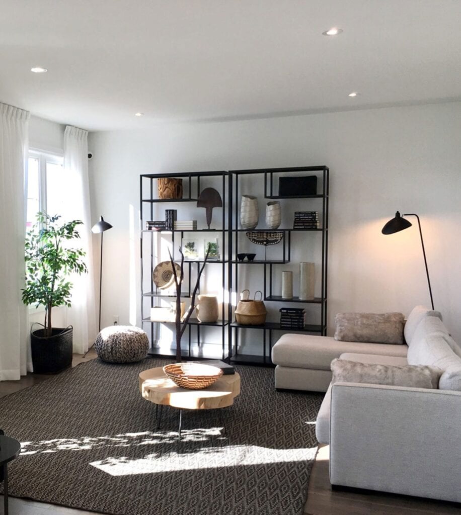 5-tendencias-para-o-inverno-de-2021-casa.com-3-my-move Sala de estar com tapete cinza, mesa de centro e sofá na cor branca e estante com diversos itens decorativos. Uma luminária de chão, junto com uma janela ampla na parede de frente, garante a iluminação do ambiente