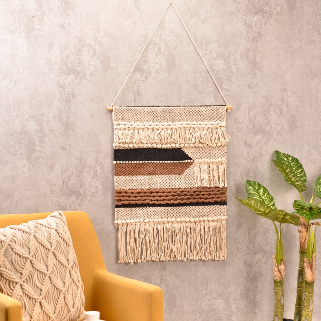 Parede de cimento queimado, com macramê branco, com detalhes em marrom e bege, uma poltrona amarela com almofada com capa de crochê e planta ao lado