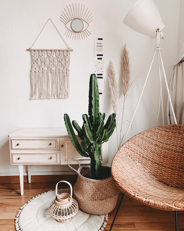 veja-solucoes-para-decorar-as-paredes-da-sua-casa-casa.com-7-unsplash-alyssa-strohmann Canto de um cômodo com cadeira pendurada no teto, um cacto grande dentro de um cesto de palha, sobre um tapete de crochê. Encostado na parede, um móvel rústico, branco e pendurado, uma peça de macramê branca