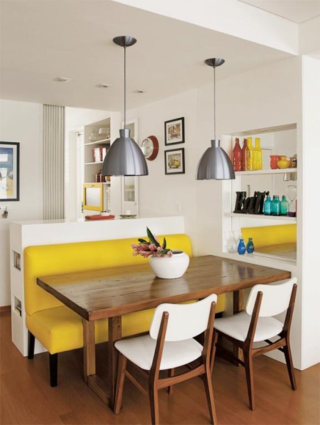 Nesta área de refeições, um sofá amarelo de couro sintético é o destaque da decoração. Uma mesa de cadeira maciça é servida por cadeiras, que acrescenta branco à combinação quente. Uma estante embutida, com fundo espelhado, amplia visualmente este canto.