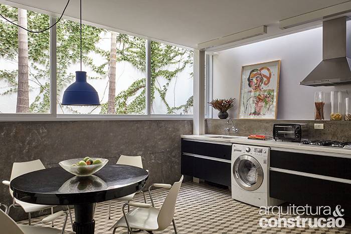 Note que a cozinha com copa inclui uma &aacute;rea de lavanderia, onde uma lavadora intercala-se entre os arm&aacute;rios de laminado preto (Formaplas). Apesar de os ambientes ficarem semienterrados, n&atilde;o s&atilde;o escuros, j&aacute; que o janel&atilde;o de alum&iacute;nio (Kiko Esquadrias) vai de uma lateral &agrave; outra, fixadoacima do barrado revestido de cimento queimado. Feita de concreto, essa base denota o refor&ccedil;o nos alicerces.