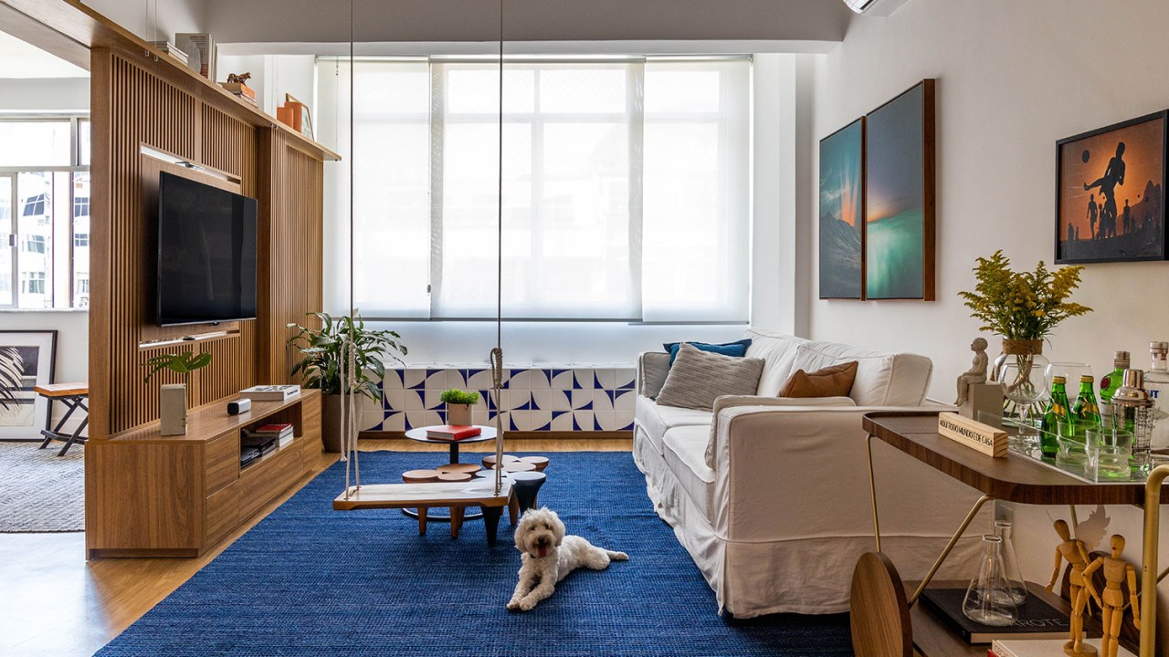 Sala de estar com tapete azul, TV sobre um painel ripado, sofá branco e tapete azul, com uma cachorrinha branca deitada sobre ele