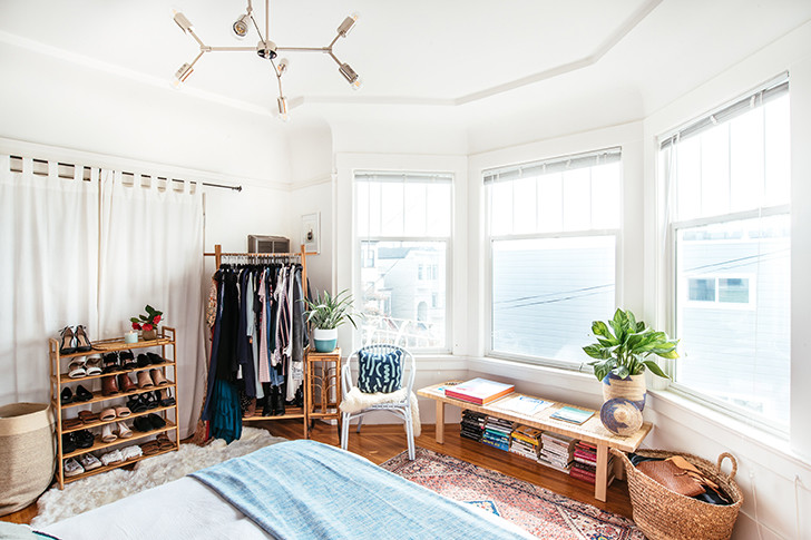 Quarto com uma parede formada por três janelas, banco de madeira - embaixo estão posicionados livros - e um guarda-roupa e sapateira abertas