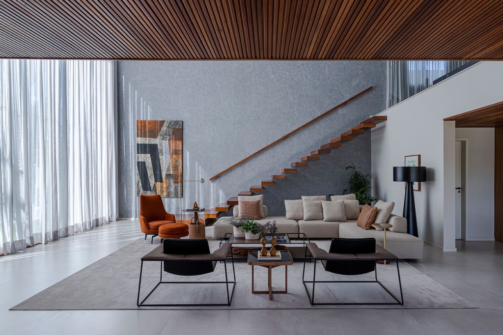estilo-contemporaneo-guia-construcao-da-casa-muxarabi-na-bahia-casa.com-sidney-quintela-gabriela-daltro-1 Sala de estar, com piso de cimento queimado, mobiliário em creme, poltrona laranja e cadeiras de couro. O teto é de maidera, e ao fundo é possível ver uma escada com degraus no mesmo material, sem guarda-corpo
