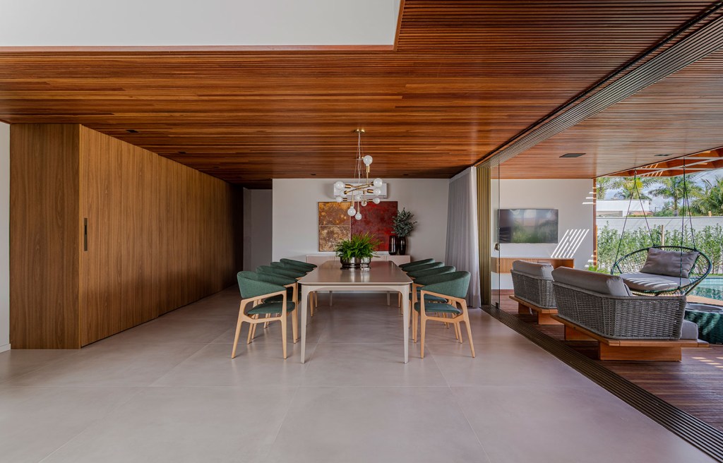 Sala de jantar, com parede e teto de amdeira, e piso de cimento queimado. A mesa de oito lugares é cinza e tem as cadeira com estofado verde