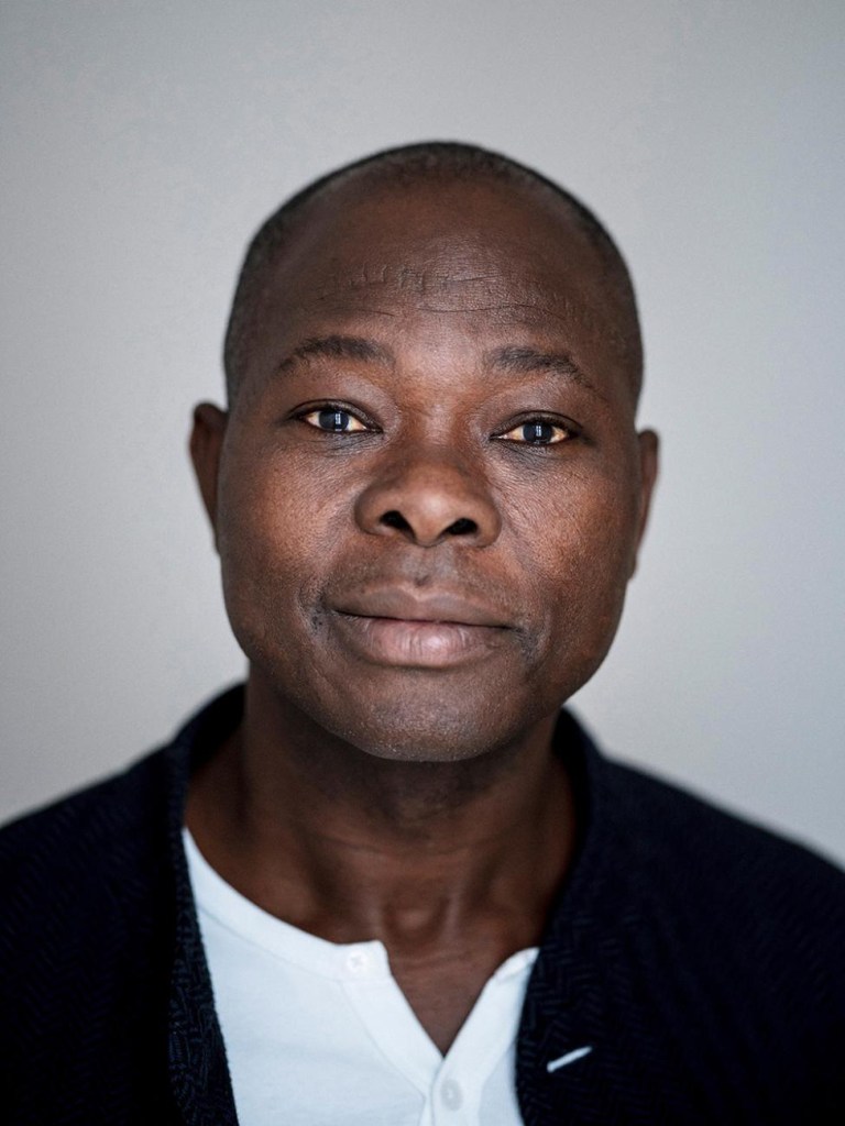 francis-kere-vencedor-pritzker-2022-lars-borges-retrato-arquiteto-kere Imagem mostra retrato do arquiteto Francis Kéré. Ele é um homem negro vestindo camisa branca sob paletó preto.