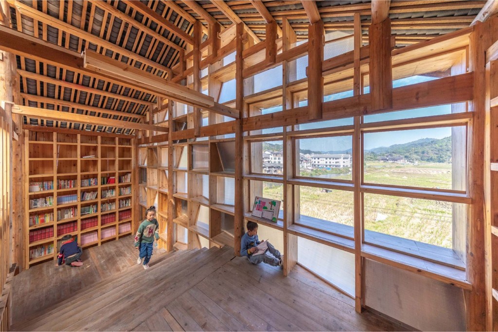 matriz-estantes-compoe-fachada-luminosa-china-sai-zhao-biblioteca-area-interna-estantes Área interna da biblioteca comunitária construída em madeira. Duas crianças brincam perto da fachada em madeira e policarbonato.