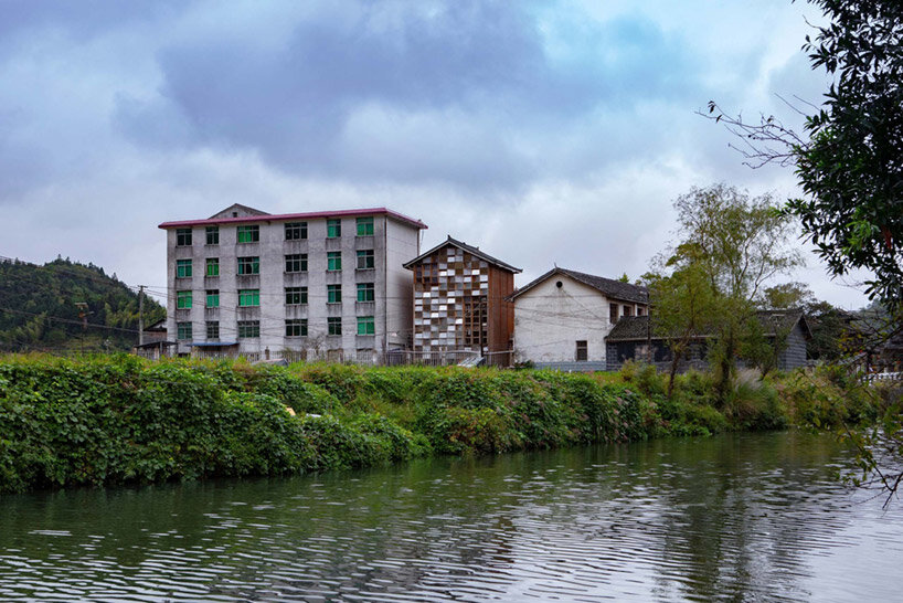 matriz-estantes-compoe-fachada-luminosa-china-sai-zhao-biblioteca-fachada-rio Vista ao longe mostra fachada de biblioteca cercada por outras duas construções, às margens de um rio.
