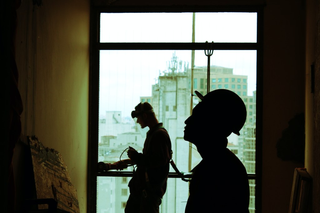 reforma-5-motivos-para-investir-projeto-arquitetonico-guilherme-cunha-trabalhadores-na-obra-capacete-de-obra Dois trabalhadores da construção civil em contraluz próximos a uma janela. Um observa a parede e o outro usa uma máquina na esquadria da janela.