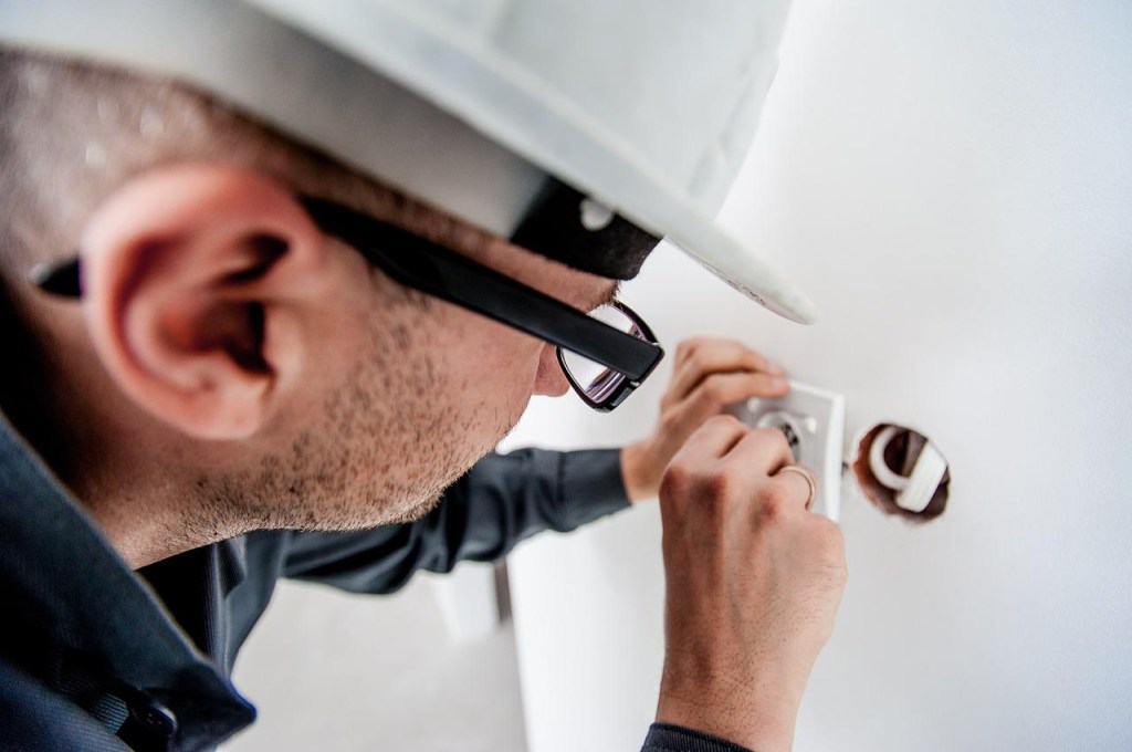 reforma-5-motivos-para-investir-projeto-arquitetonico-michal-jarmoluk-eletricista-tomada-capacete-obra Homem com capacete de obra e óculos mexendo em uma tomada perto da parede.