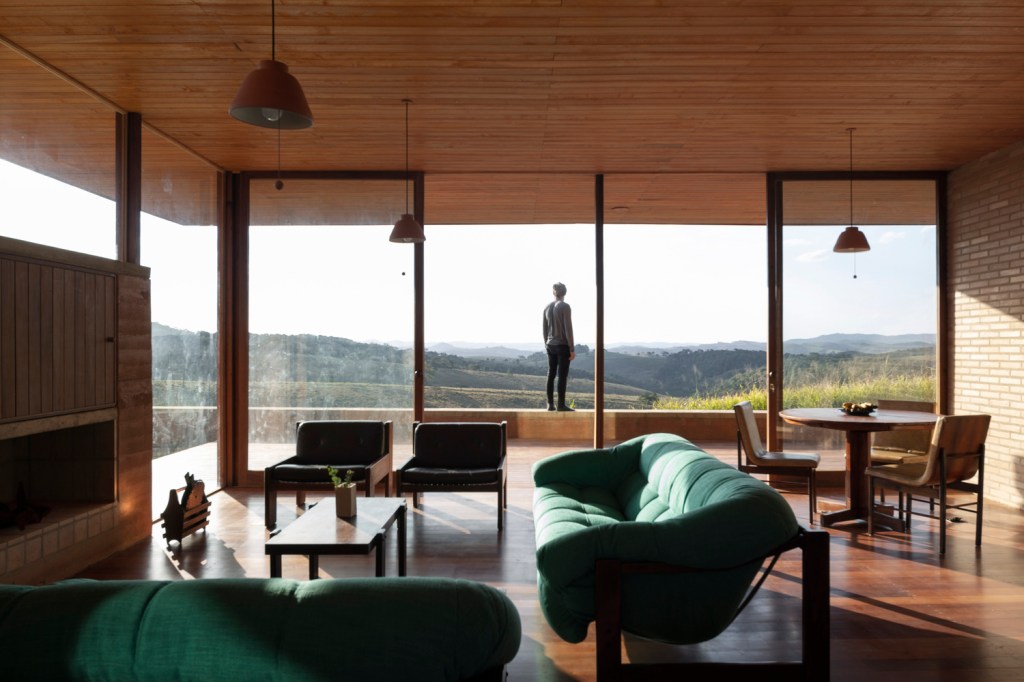 Sala de estar em casa de campo com grandes aberturas de vidro que permitem ver pessoa na varanda diante da paisagem montanhosa.