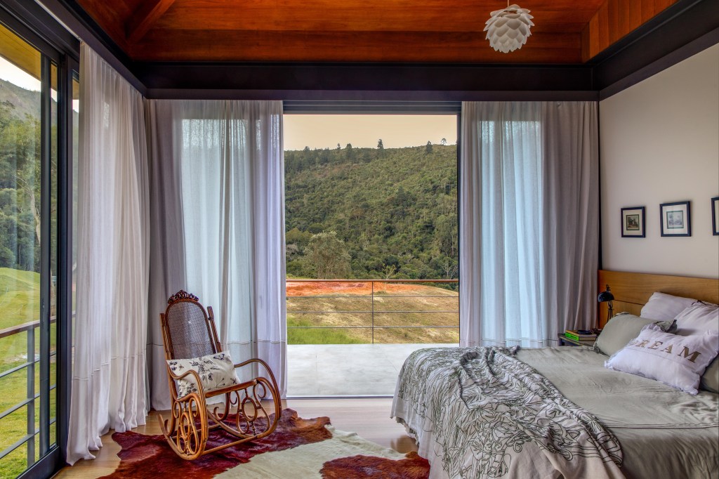 17-casa-de-campo-mistura-elementos-tradicionais-e-contemporaneos Quarto com cama de casal e cadeira de balanço de madeira com vista para serra