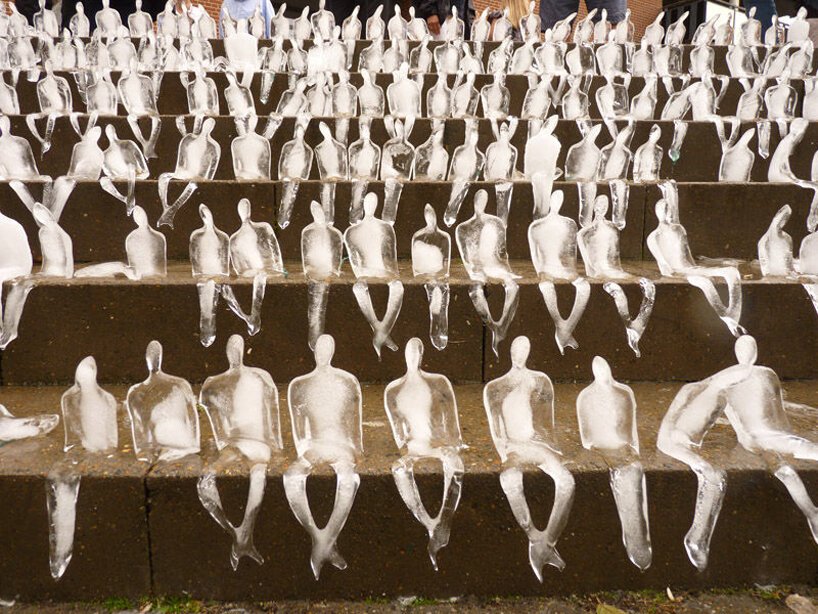 Pequenas esculturas de gelo derretendo de pessoas sentadas uma ao lado da outra de cabeças levemente baixas numa escadaria.