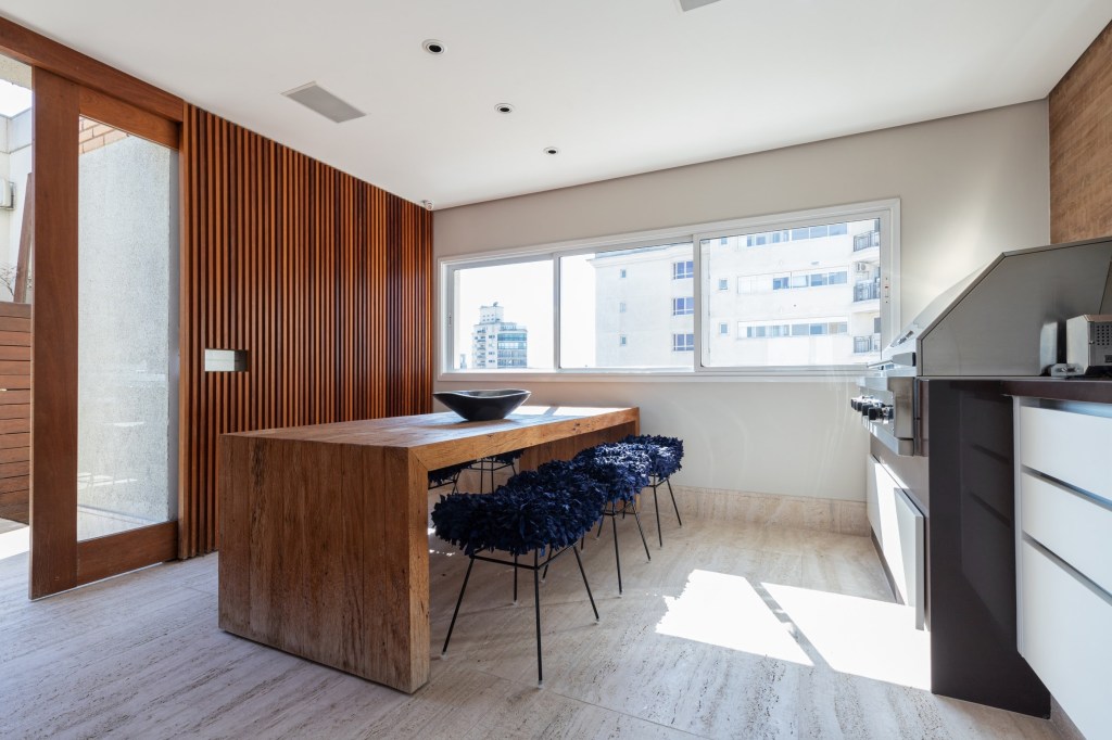 Foto mostra cozinha com mesa de madeira e banquetas em tecido azul marinho. Na lateral esquerda vê-se um painel ripado de madeira que 'esconde' a porta de correr, também de madeira.