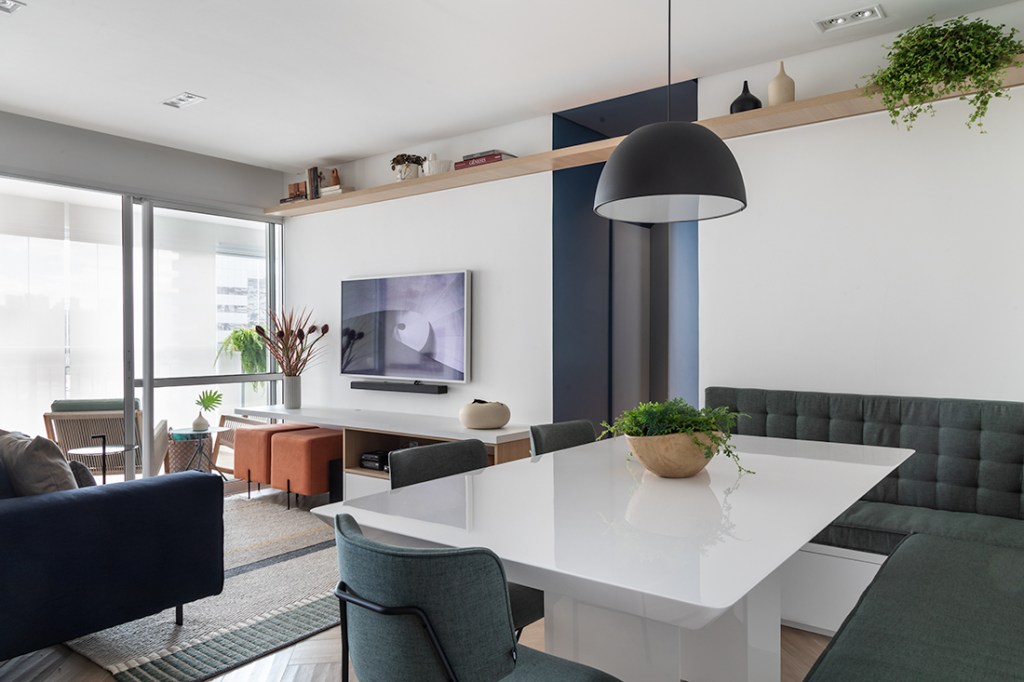 o-que-considerar-antes-comprar-apartamento-evelyn-müller-estar-integrado-tons-de-cinza-pendente-industrial-prateleira-alta Mesa de jantar em laca branca com banco cadeiras em tom de cinza escuro e, ao fundo, sala de estar com TV e vista para varanda.