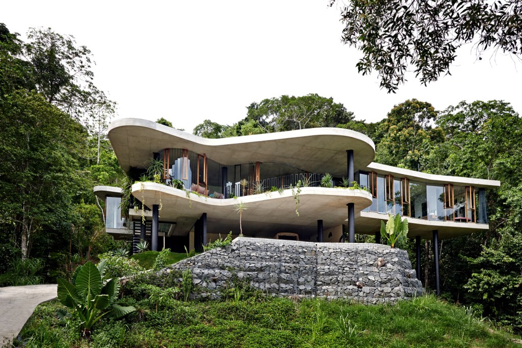 Fachada de casa suspensa por pilares de concreto pintados de preto e piso e cobertura em concreto com linhas curvas, cercada por árvores e vegetação arbustiva.