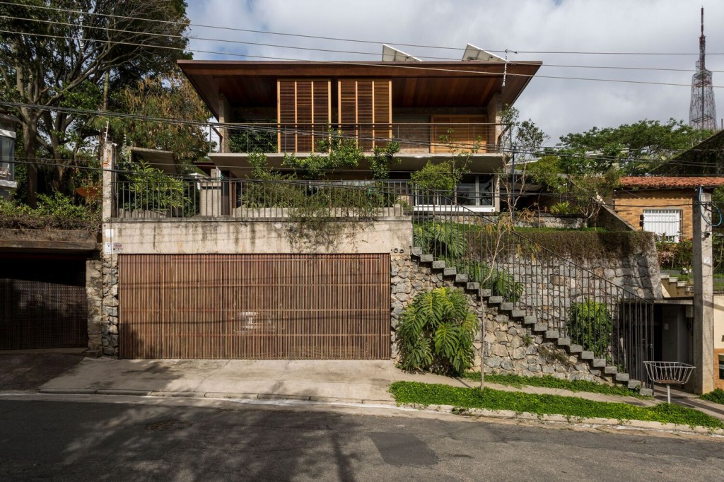 Foto mostra fachada frontal da casa, com garagem na divisa da calçada e escada de acesso ao piso superior, com a construção principal ao fundo.