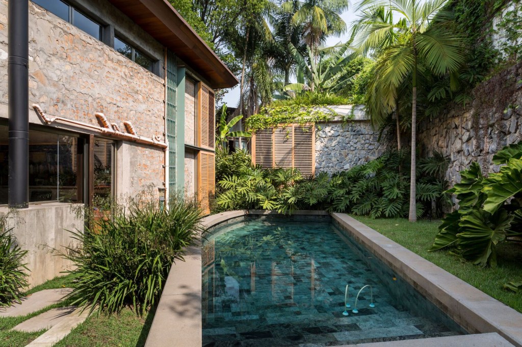 Foto mostra piscina cercada de jardim com fachada lateral da casa feita com paredes de entulho.