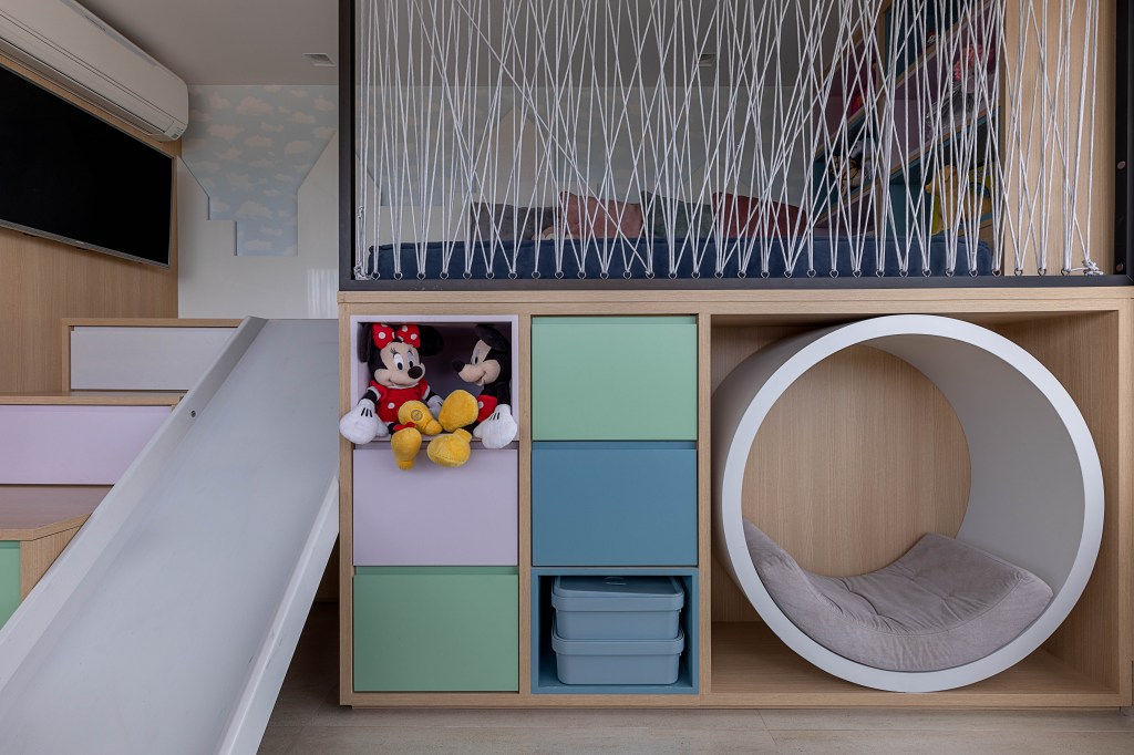 Na premissa de criar um ambiente divertido, lúdico e organizado, a designer de interiores Norah Carneiro desenvolveu um espaço kids com caixas organizadoras, gavetas coloridas na escada e, para garantir a diversão, foi atribuído em meio a marcenaria personalizada um escorregador. Na parte alta da estrutura, a profissional inseriu papel de parede com céu azul e um recorte em relevo de castelos, além de outros nichos para organizar os brinquedos.