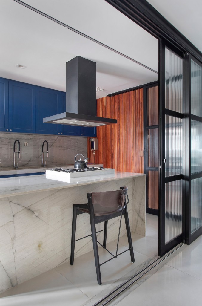 ape-160m-paineis-madeira-ripada-sofa-verde-design-nacional-roby-studio-arquitetura-03-cozinha Cozinha com ilha revestida de mármore e marcenaria azul; porta de correr de vidro.