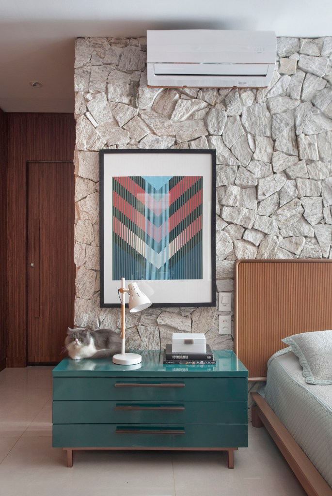 ape-160m-paineis-madeira-ripada-sofa-verde-design-nacional-roby-studio-arquitetura-11-quarto-parede-pedra Quarto com cama, parede de pedra, cabeceira de madeira e mesa lateral verde.