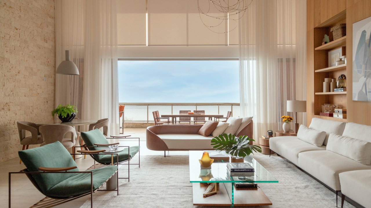 Sala de estar com pé-direito alto, sofás brancos, tapete claro, mesa de centro com tampo de vidro e poltronas verdes, vista para varanda.