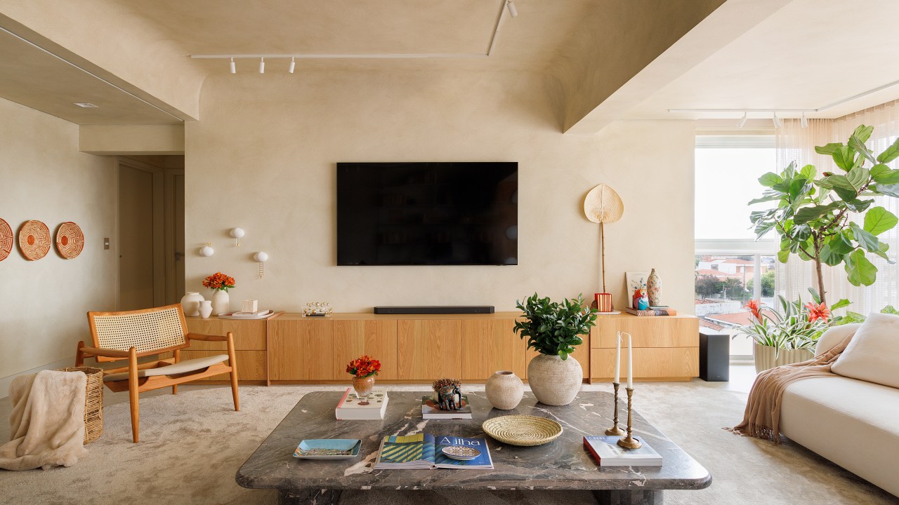 Sala de tv em tons terrosos claros com mesa de centro, sofá branco e rack de madeira.