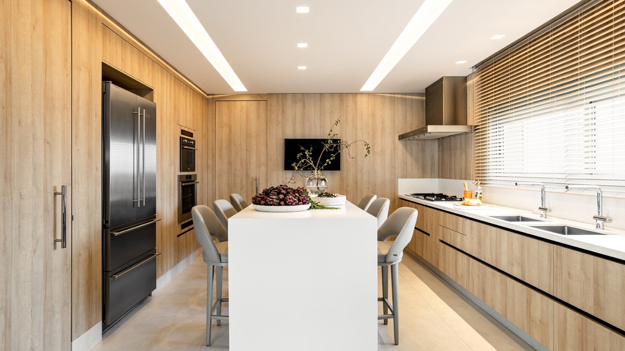 Cozinha layout clean elegante revestimento amadeirado Mota Arquitetura ilha bancada cadeiras tv torre quente pia cortina