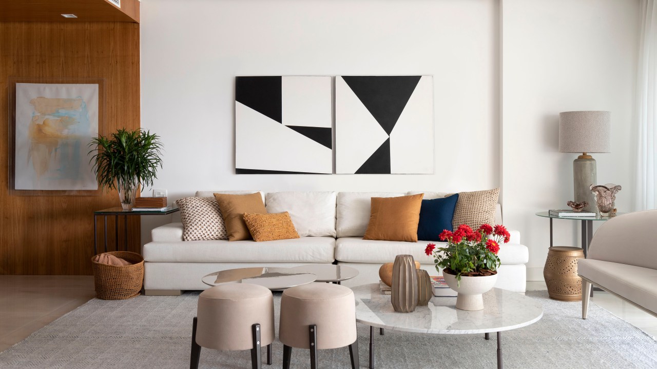 Sala de estar com sofá branco, almofadas em tons terrosos, quadro em cima do sofá, pufes e mesa de centro.