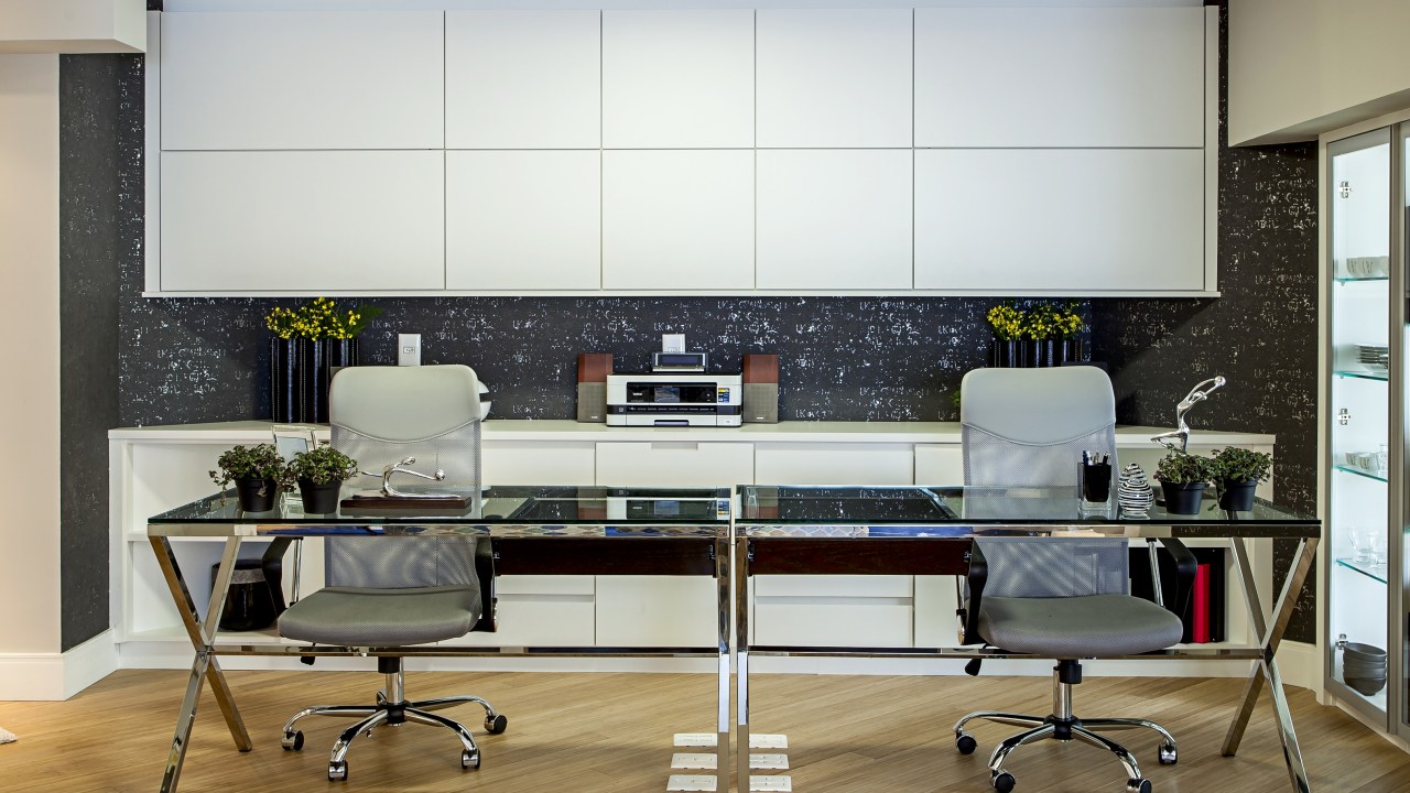 Home office para duas pessoas com marcenaria branca e mesa com tampo de vidro.