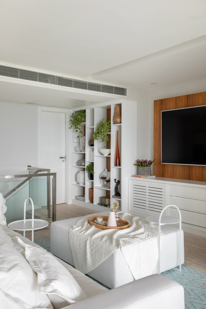 sem-demolicao-ape-280m-pecas-design-sala-intima-bianca-da-hora-09 Sala íntima com sofá e pufe brancos, painel de tv e estante com nichos brancos.