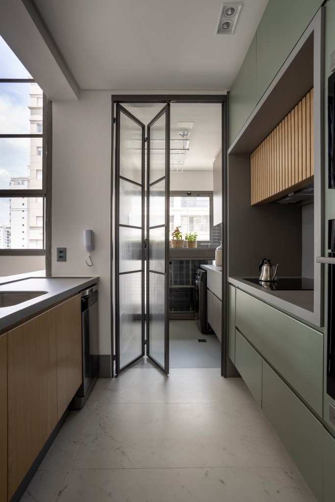 Apê de 110 m² com pé-direito duplo vira duplex com mezanino metálico Apê de 110 m² com pé-direito duplo vira duplex com mezanino metálico. NA foto cozinha separada da lavanderia por uma porta camarao de vidro canelado.