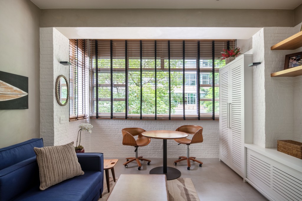 ape-90m-cozinha-com-azulejos-azuis-studio021arquitetura-13-varnda Varanda com parede de tijolinhos brancos, mesa de madeira redonda pequena e duas cadeias. Sala de estar com sofá azul.