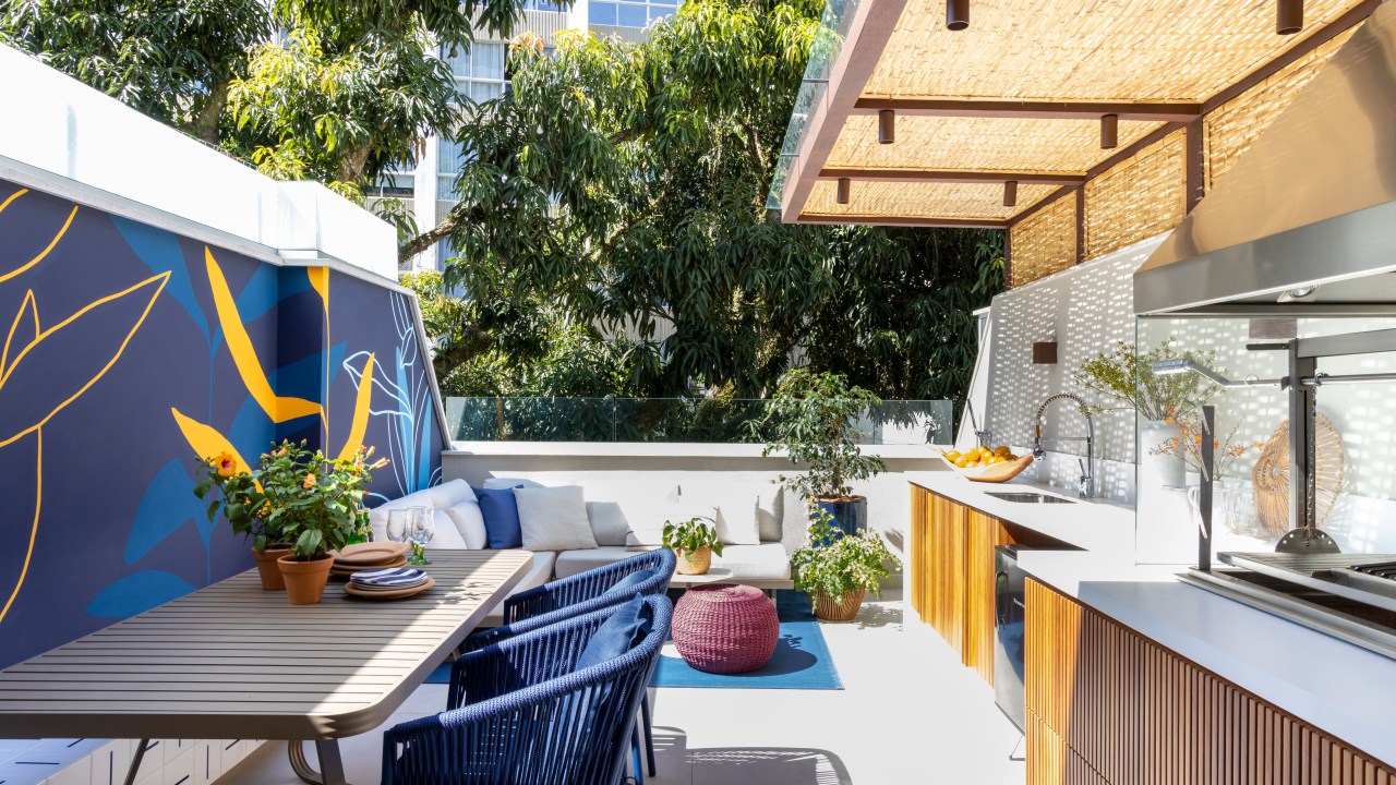 Terraço com mesa de madeira, cadeiras azuis, sofá branco, churrasqueira, bancada branca.