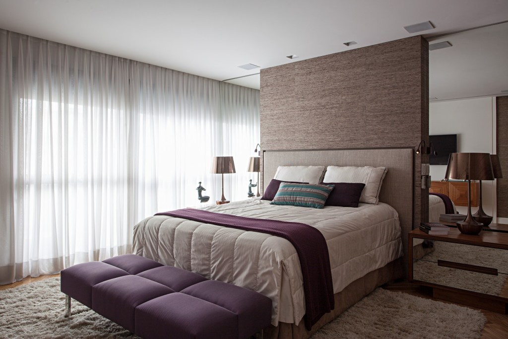 como-escolher-o-tapete-certo-korman-arquitetos-04 Quarto com tapete felpudo, cama de casal e banco roxo.