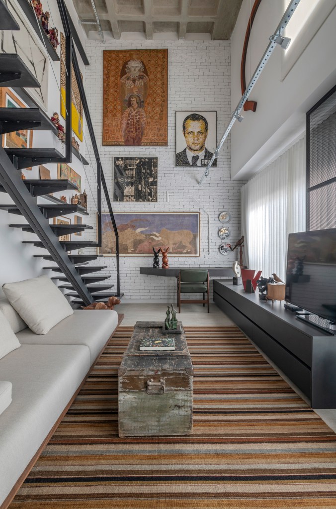 loft-duplex-compacto-64m-decor-industrial-arte-piacesi-arquitetos-10 Sala de estar com sofá branco, tapete listrado, baú rústico como mesa de centro e rack com tv; gallery wall ao fundo.