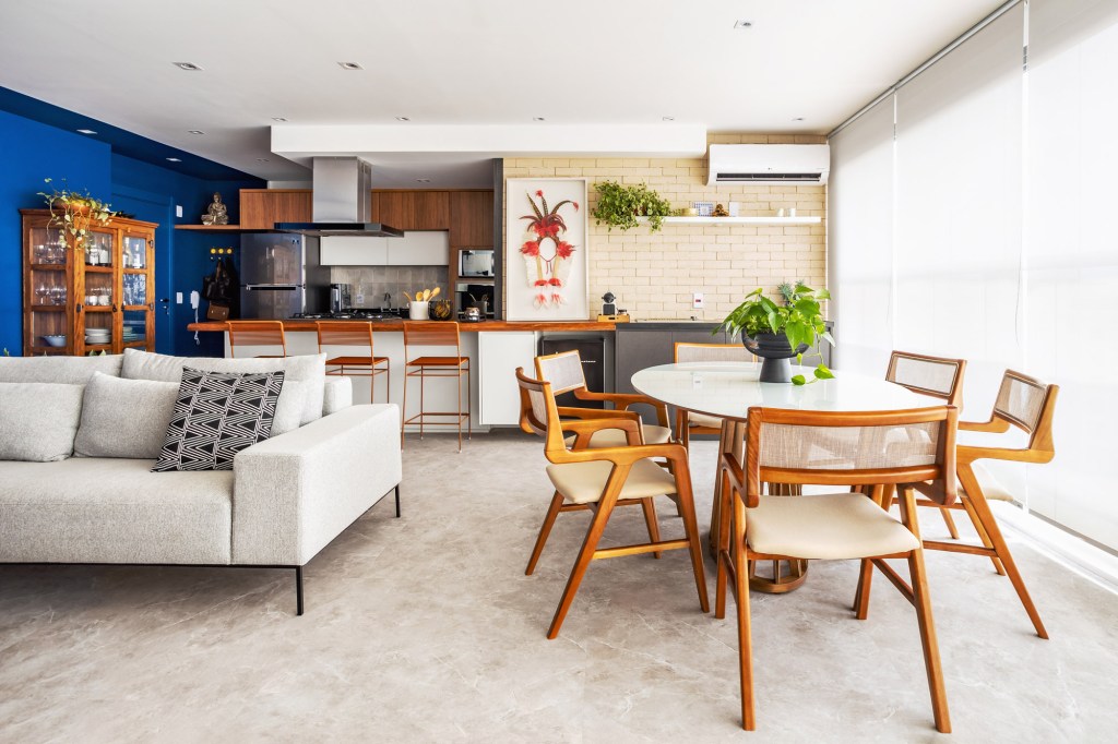 Sala integrada com varanda e cozinha; piso vinílico marmorizado, mesa oval branca, sofá branco e parede de tijolinhos.