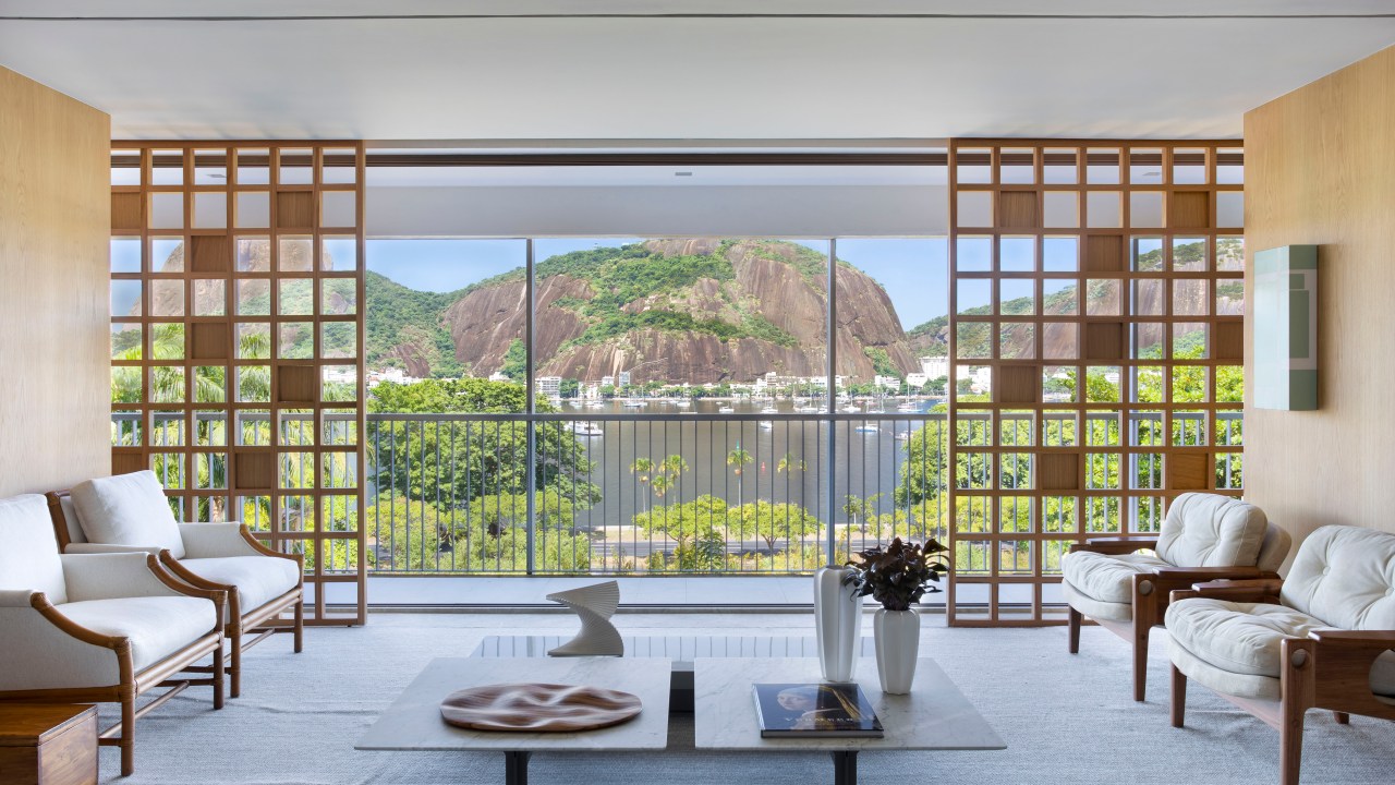 Sala de estar com divisórias de muxarabi, sofá cinza claro, poltrona brancas e vista para paisagem.