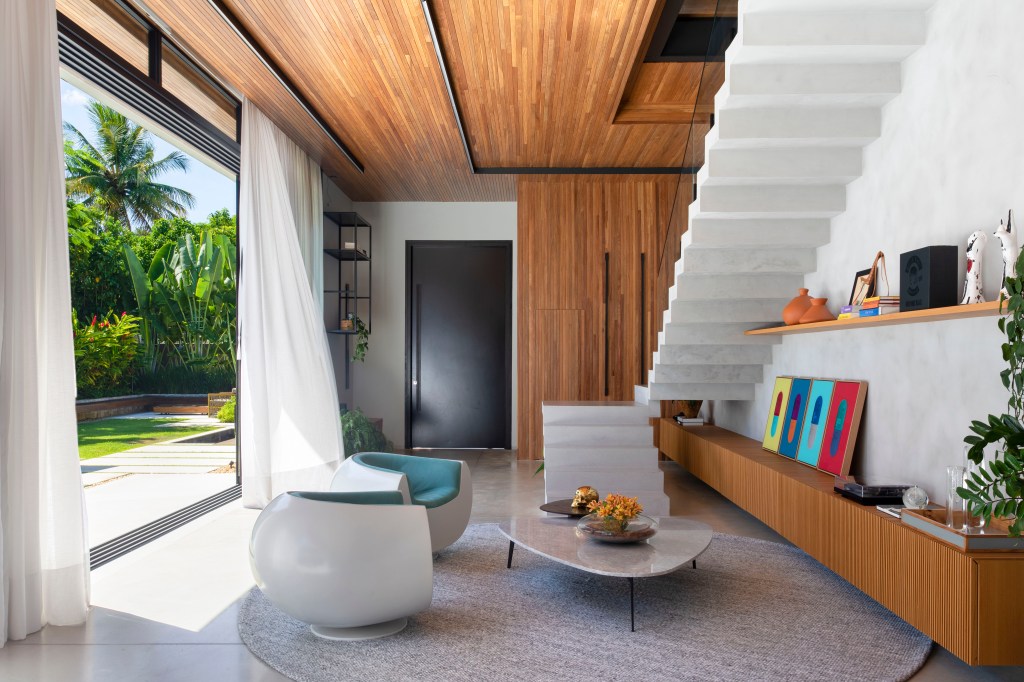 pe-direito-alto-teto-ripado-casa-634m-up3-arquitetura-06 Sala de estar com poltronas, cortina branca, escada branca e teto ripado.